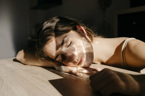 Portrait of young woman with eyes closed leaning on wooden table at sunlight - TCEF00035