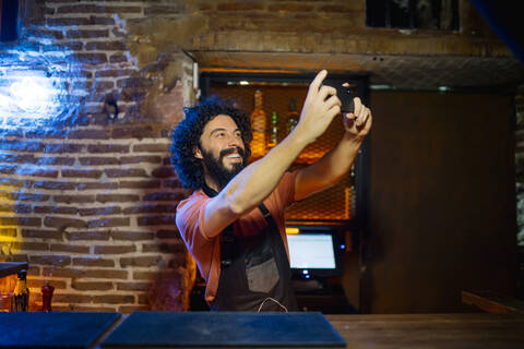 Junger Mann, der in einer Bar arbeitet und ein Selfie mit seinem Smartphone macht, lizenzfreies Stockfoto