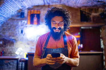 Junger Mann, der in einer Bar arbeitet und ein Smartphone benutzt - CJMF00242