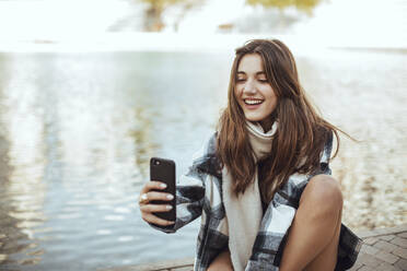 Young brunette woman using smartphone and taking a selfie at lake - ACPF00659