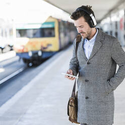 Junger Geschäftsmann mit Mobiltelefon und Kopfhörern am Bahnhof - UUF20147