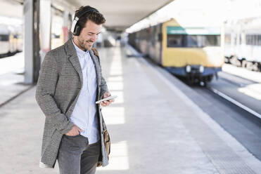 Junger Geschäftsmann mit Mobiltelefon und Kopfhörern am Bahnhof - UUF20148