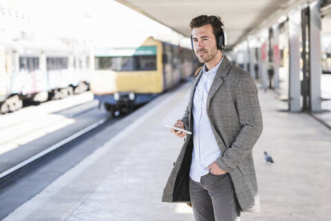 Junger Geschäftsmann mit Mobiltelefon und Kopfhörern am Bahnhof, lizenzfreies Stockfoto