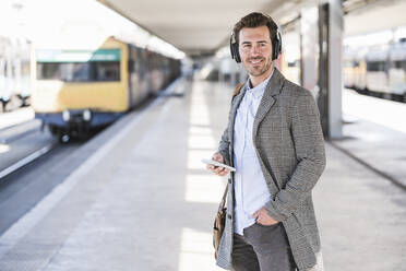 Lächelnder junger Geschäftsmann mit Mobiltelefon und Kopfhörern auf dem Bahnhof - UUF20150