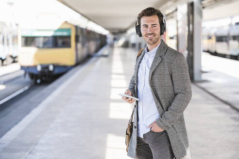 Smiling young businessman with cell phone and headphones at the train station - UUF20150