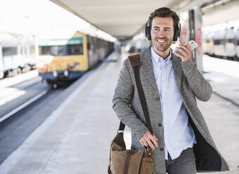Lächelnder junger Geschäftsmann mit Mobiltelefon und Kopfhörern auf dem Bahnhof - UUF20151