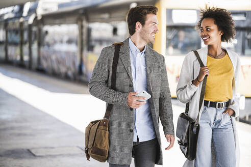 Happy young businessman and businesswoman walking and talking at the train station - UUF20154