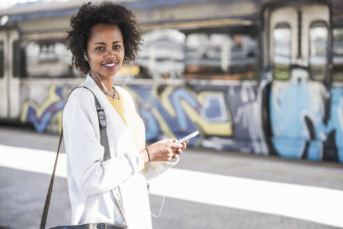 Portrait of smiling young woman with cell phone and earphones at the train station - UUF20162