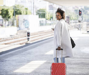 Smiling young woman with rolling suitcase at the train station - UUF20168
