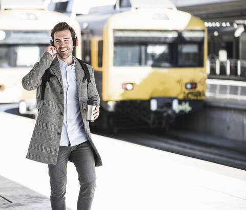 Glücklicher junger Geschäftsmann mit Kaffeebecher und Kopfhörern beim Spaziergang am Bahnhof, lizenzfreies Stockfoto