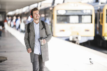 Junger Geschäftsmann mit Kaffeebecher und Kopfhörer am Bahnhof - UUF20171