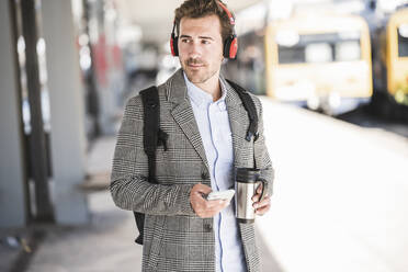 Junger Geschäftsmann mit Mobiltelefon und Kopfhörern am Bahnhof - UUF20173