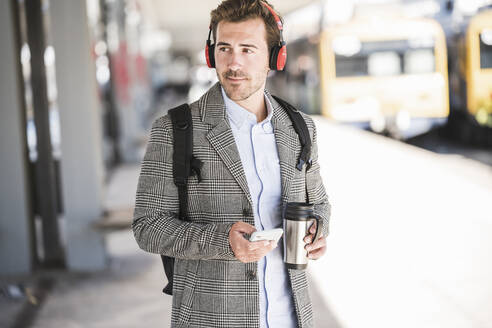 Young businessman with cell phone and headphones at the train station - UUF20173