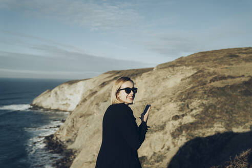 Rückansicht einer jungen blonden Frau mit Smartphone an der Küste - MTBF00308