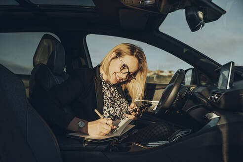 Junge blonde Frau benutzt ihr Smartphone im Auto und schreibt in ein Notizbuch - MTBF00309