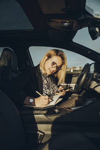 Junge blonde Frau benutzt ihr Smartphone im Auto und schreibt in ein Notizbuch, lizenzfreies Stockfoto