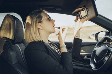 Junge blonde Geschäftsfrau in einem Auto trägt Lippenstift auf - MTBF00315