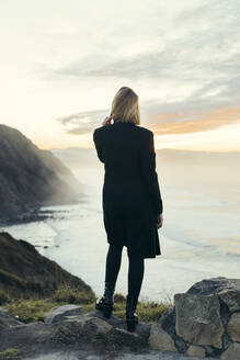 Rückansicht einer jungen blonden Frau mit Smartphone an der Küste bei Sonnenuntergang - MTBF00319