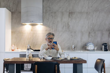 Älterer Mann sitzt am Tisch in der Küche zu Hause und benutzt ein Mobiltelefon - SODF00515