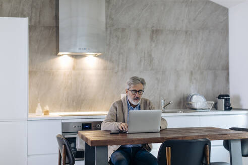 Älterer Mann mit Laptop am Tisch in der Küche zu Hause - SODF00544