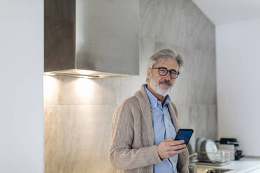 Porträt eines reifen Mannes mit Mobiltelefon in der Küche zu Hause - SODF00550
