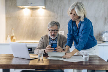 Mature couple with papers, cell phone and laptop on kitchen table at home - SODF00562