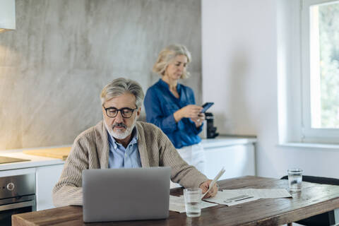 Mature man with papers and laptop on kitchen table at home with wife in background stock photo