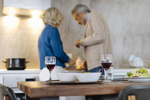 Red wine glasses on kitchen table with mature couple preparing dinner - SODF00606