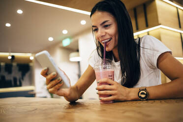 Schwarzhaarige Frau trinkt einen Smoothie und benutzt ihr Smartphone in einem Café - OYF00102