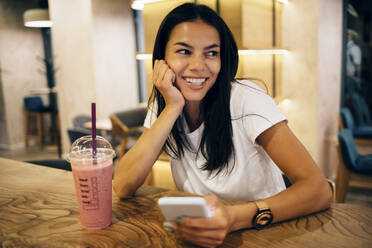 Schwarzhaarige Frau trinkt einen Smoothie und benutzt ihr Smartphone in einem Café - OYF00104