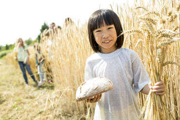 Kleines asiatisches Mädchen steht auf einem Feld und hält einen Laib Brot und Weizenähren - WESTF24456