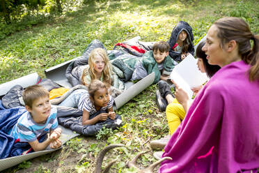Teacher reading story to school children, camping in the forest - WESTF24486