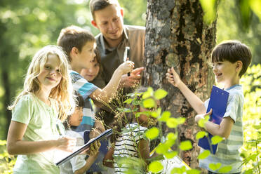 School children examining tree bark in forest with their teacher - WESTF24546