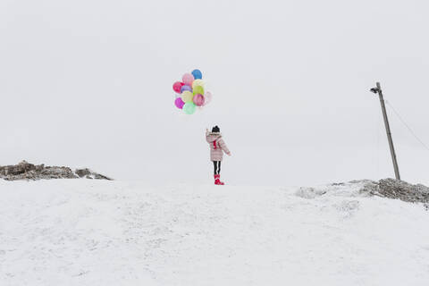 Rückenansicht eines Mädchens mit Luftballons auf einem schneebedeckten Hügel im Winter, lizenzfreies Stockfoto