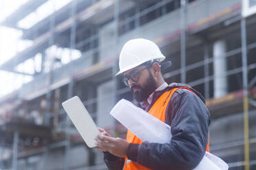 Bauingenieur mit Schutzhelm und Sicherheitsweste, der auf einer Baustelle ein Tablet benutzt - SGF02579
