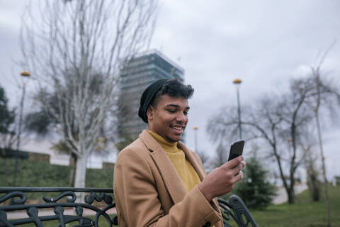 Lächelnder junger Mann sitzt auf einer Bank und benutzt ein Mobiltelefon, lizenzfreies Stockfoto