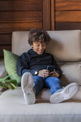 Smiling relaxed boy using cell phone on couch at home - DLTSF00399