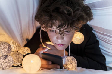 Boy with chain of lights underneath bedcover using cell phone - DLTSF00416