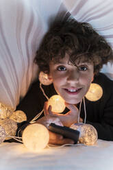Porträt eines lächelnden Jungen mit Lichterkette und Mobiltelefon unter der Bettdecke - DLTSF00417