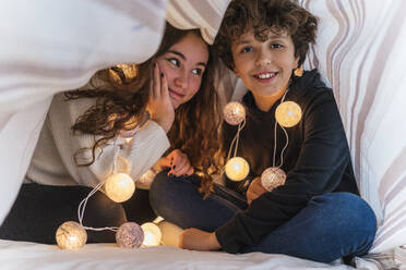 Portrait of happy brother and sister with chain of lights underneath bedcover - DLTSF00418