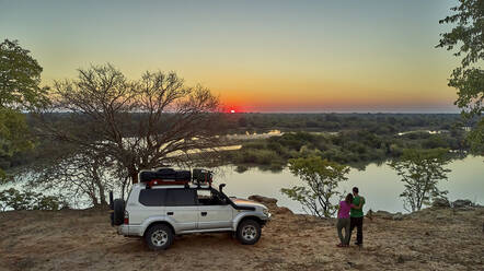 Luftaufnahme eines Paares mit ihrem Jeep, das den Sonnenuntergang vor dem Fluss genießt, Cunene-Flussgebiet, Angola - VEGF01437