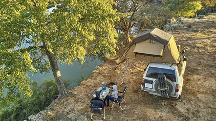 Luftaufnahme eines Paares, das mit seinem Jeep und einem Dachzelt am Flussufer des Cunene-Flusses frühstückt, Angola - VEGF01440