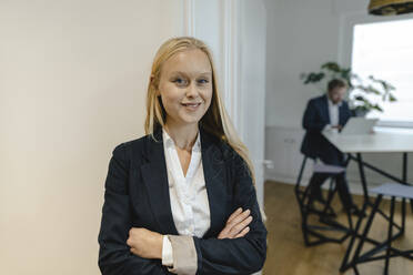 Porträt einer selbstbewussten jungen Geschäftsfrau im Büro mit einem Geschäftsmann im Hintergrund - GUSF03203
