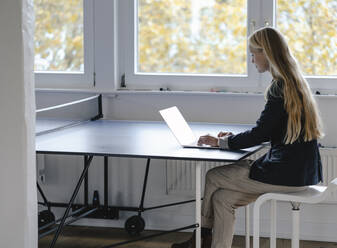 Junge Geschäftsfrau mit Laptop an der Tischtennisplatte im Büro - GUSF03323