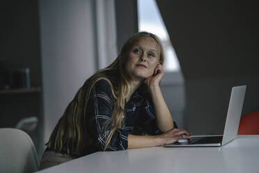 Casual young businesswoman with laptop in office looking away - GUSF03333