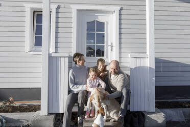 Glückliche Familie mit zwei Kindern und zwei Hunden vor ihrem Haus sitzend - KMKF01209