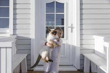 Little girl carrying cat on her arms in front of house entrance - KMKF01210