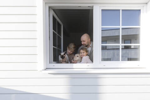 Vater mit seinen beiden Kindern, Hund und Katze am geöffneten Fenster ihres Hauses, lizenzfreies Stockfoto