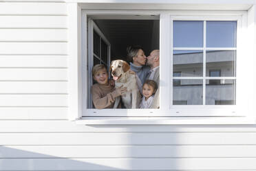 Glückliche Familie mit zwei Kindern und einem Hund am geöffneten Fenster ihres Hauses - KMKF01213