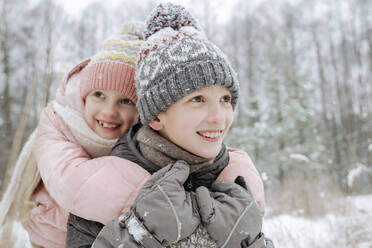 Porträt eines Jungen, der seine kleine Schwester im Winterwald huckepack nimmt - EYAF00845